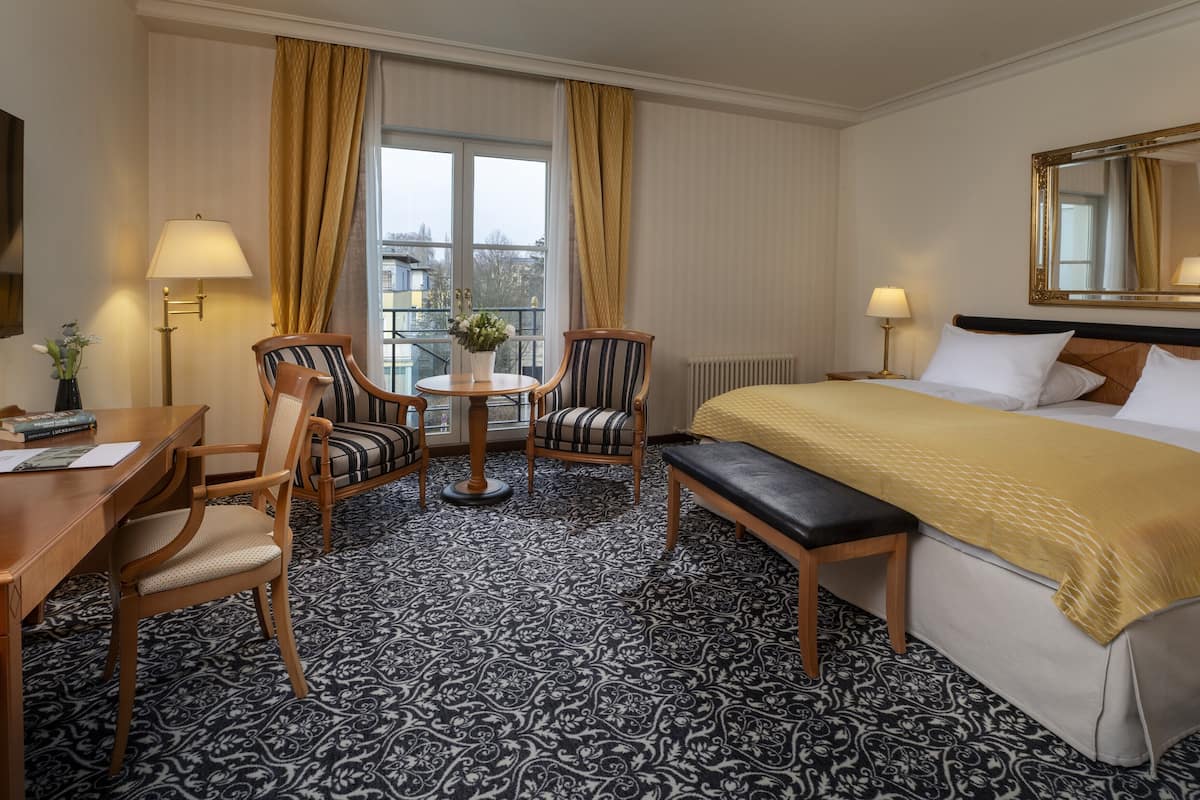 Habitación de hotel con cama, escritorio, dos sillas, una mesita con un florero y vistas a la ciudad a través de amplios ventanales.