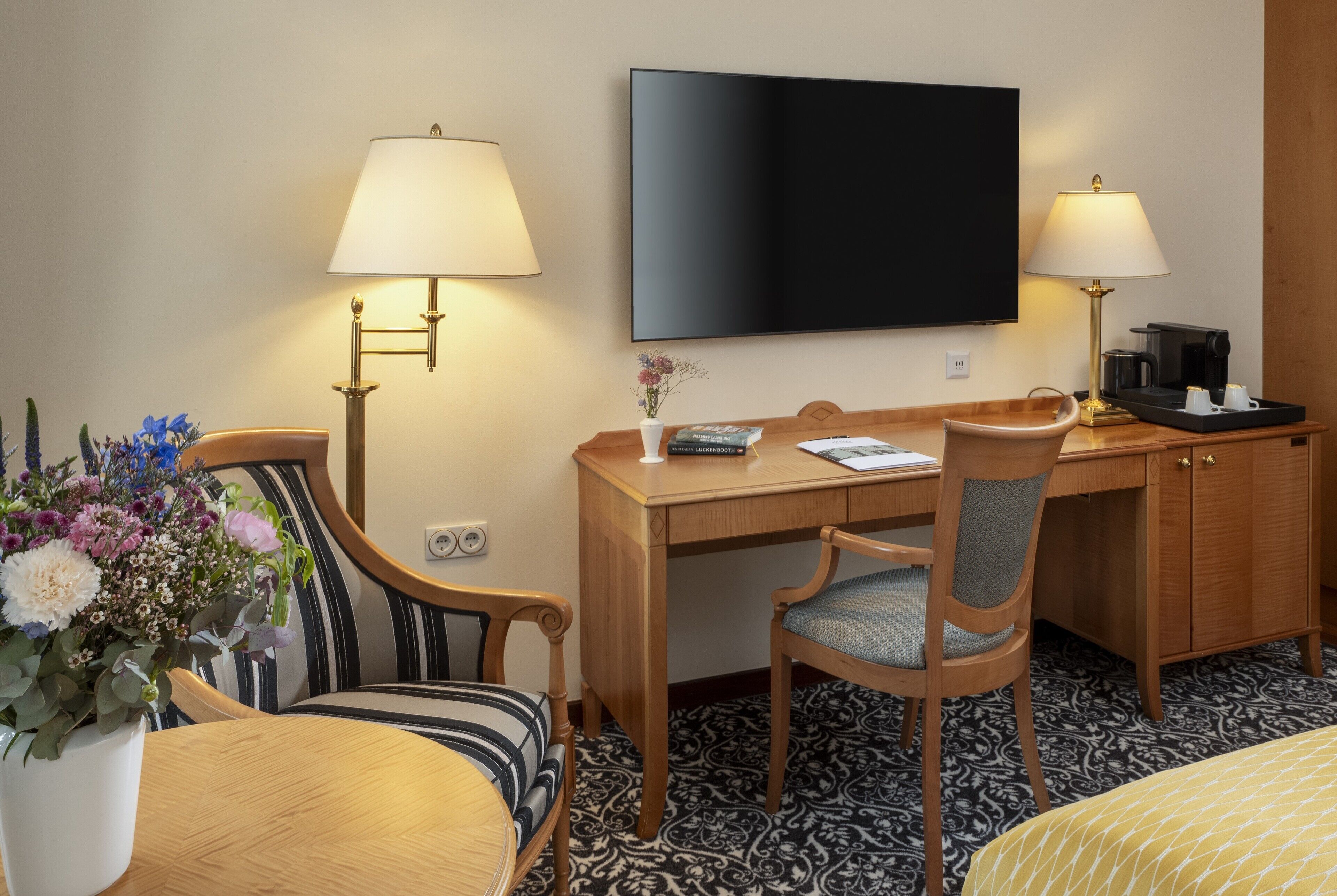 A hotel room with a wooden desk, two chairs, a television, a coffee maker, and a flower vase.