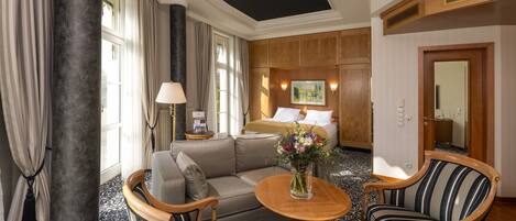 A hotel room with a bed, a sofa, two armchairs, a small table with a vase of flowers, a lamp, and a painting on the wall.