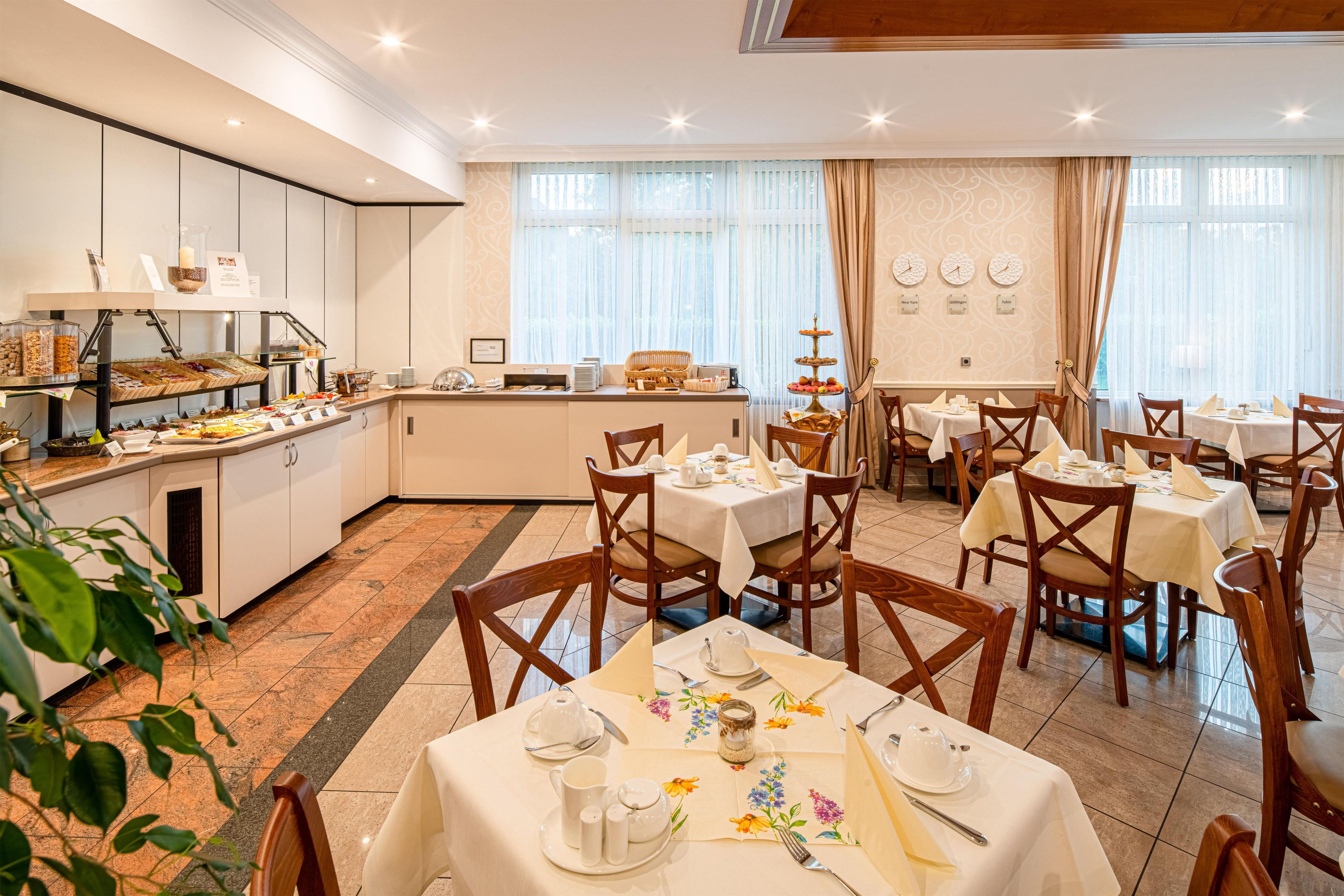 Un comedor de hotel con varias mesas dispuestas para el desayuno, un bufé con distintos alimentos y un reloj en la pared que muestra diferentes husos horarios.