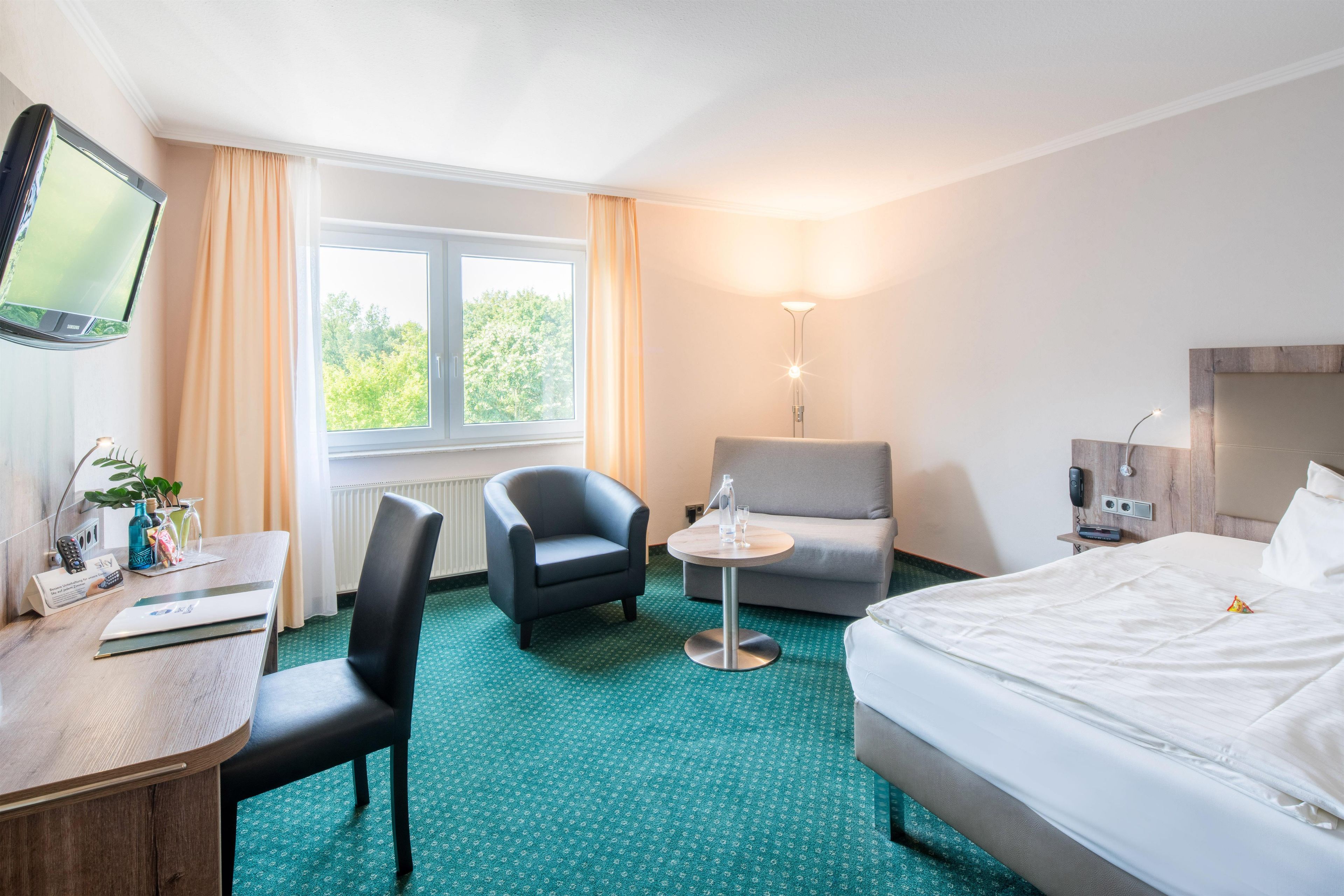 A hotel room with a bed, a desk with a computer, a chair, a sofa, a TV, and a window with curtains.