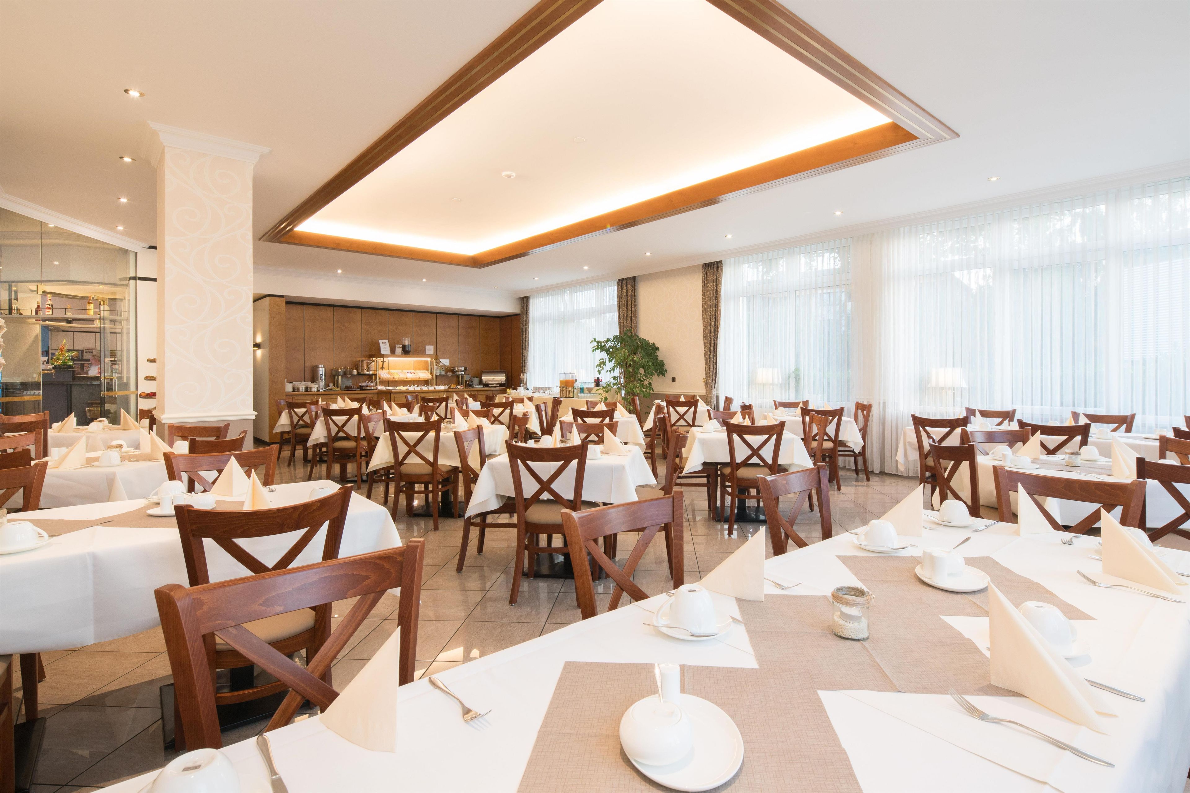 A spacious dining area with multiple tables and chairs, a kitchen in the background, and large windows allowing natural light.