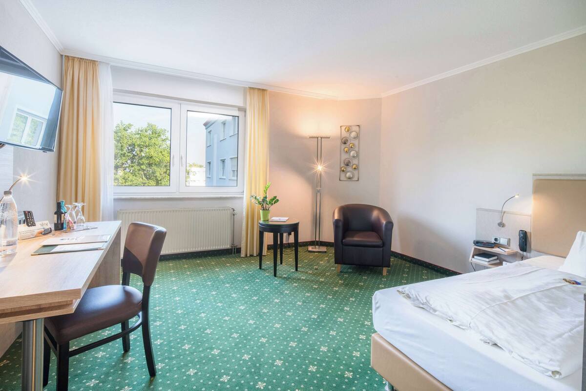 A hotel room with a bed, a desk, a chair, a small table, a lamp, and a window with curtains.