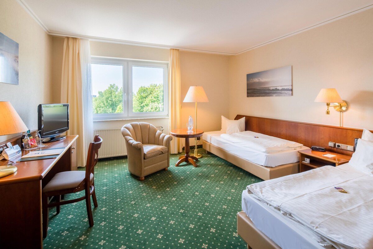 A hotel room with a desk, chair, two beds, a lamp, and a window with a view of trees.