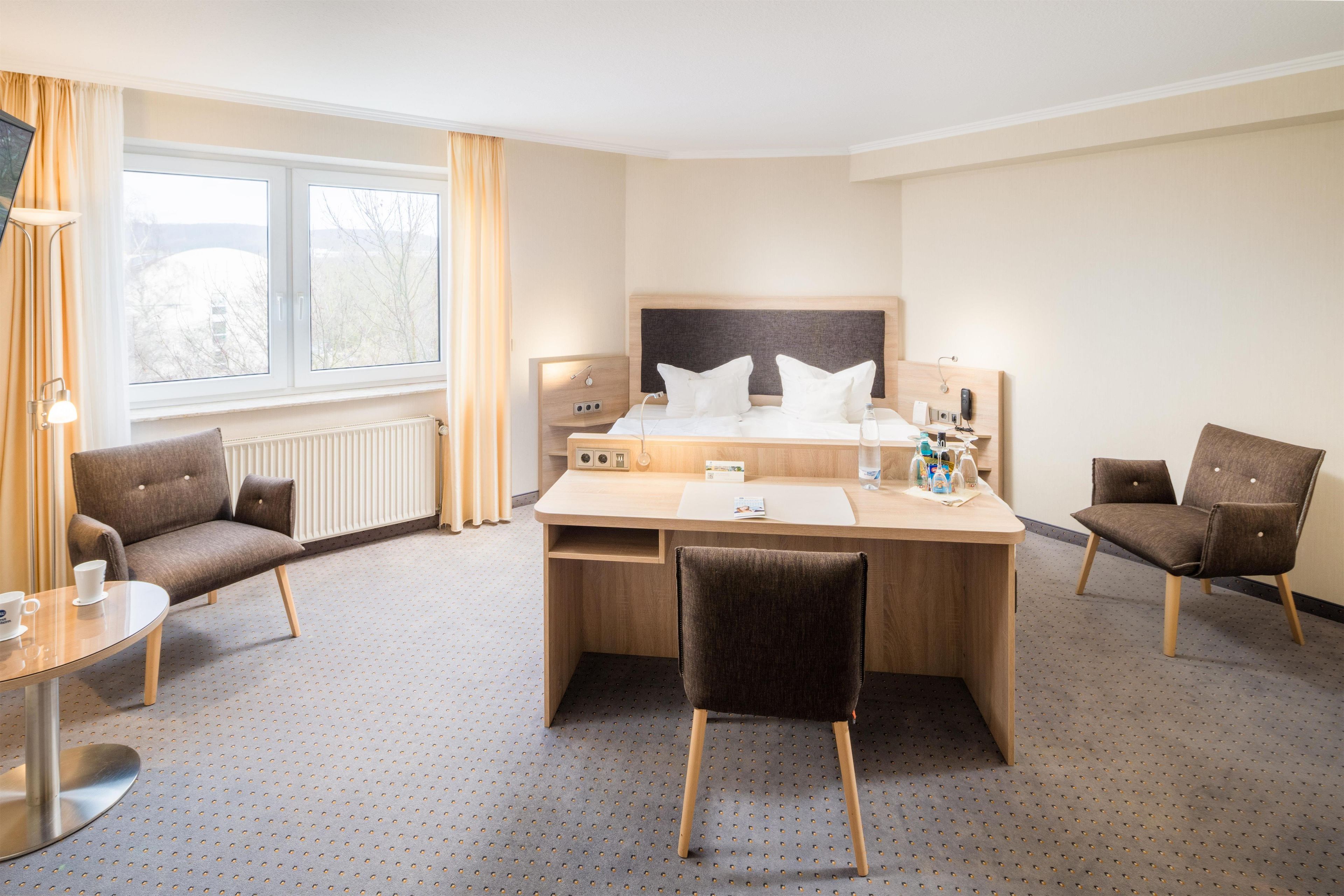 A hotel room with a bed, two armchairs, a desk, a window with curtains, and a radiator.