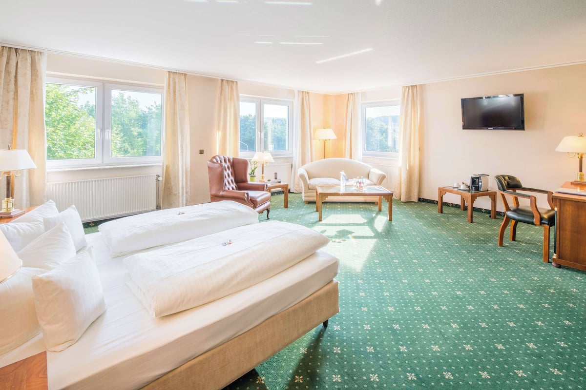 A hotel room with a large bed, two armchairs, a small table, a TV, and a window with curtains.