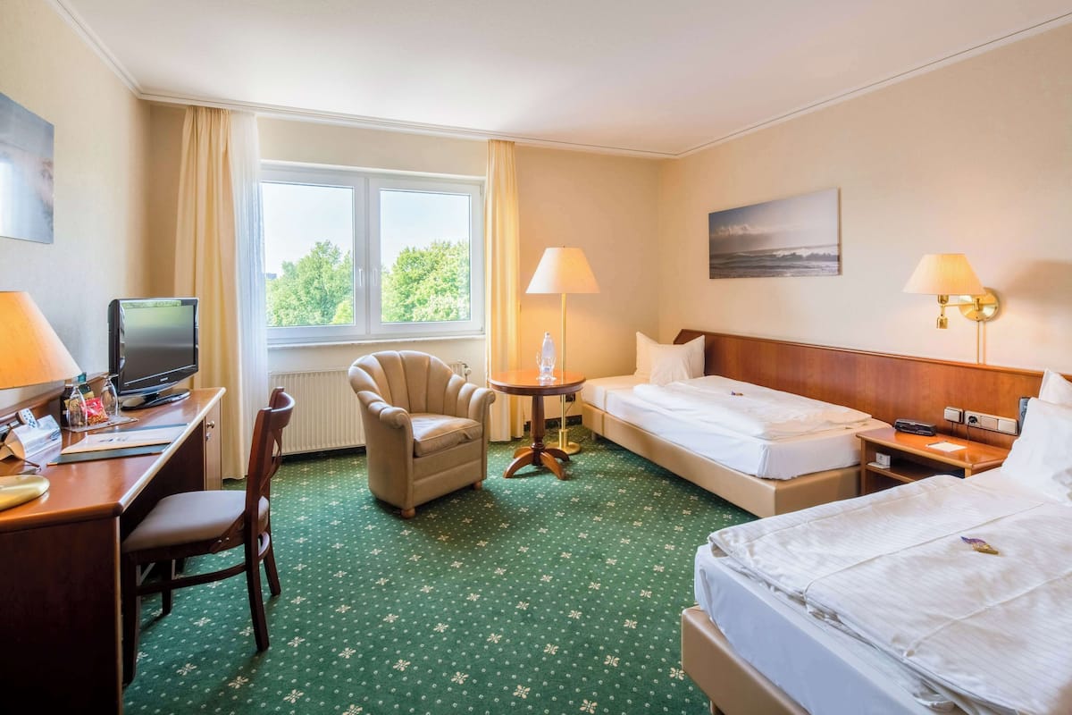 A hotel room with a desk, chair, two beds, a small table, lamps, and a window with curtains.