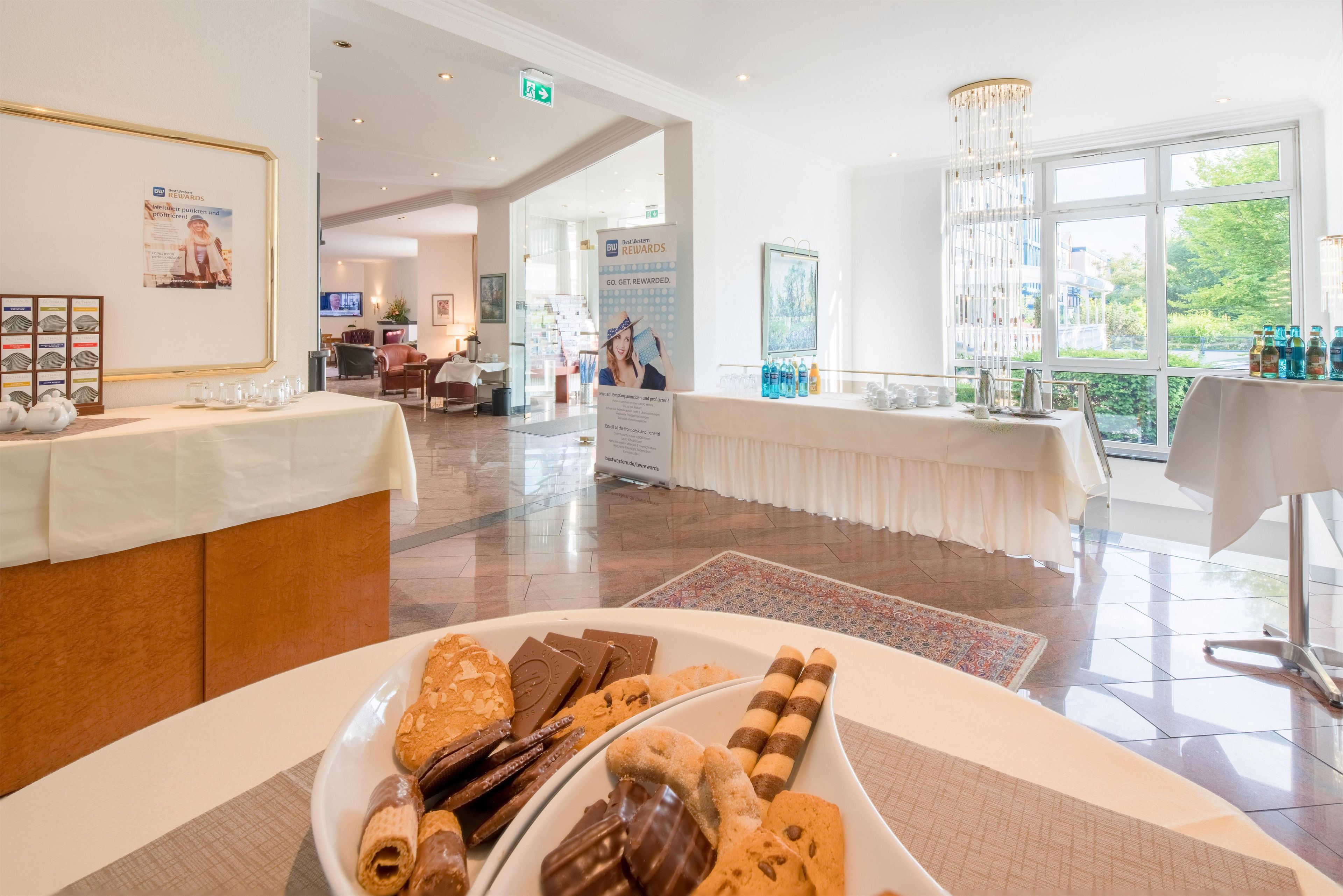 Et hotel lobby med en receptionsdisk, et buffetbord med bagværk og udsigt til det fri gennem store vinduer.