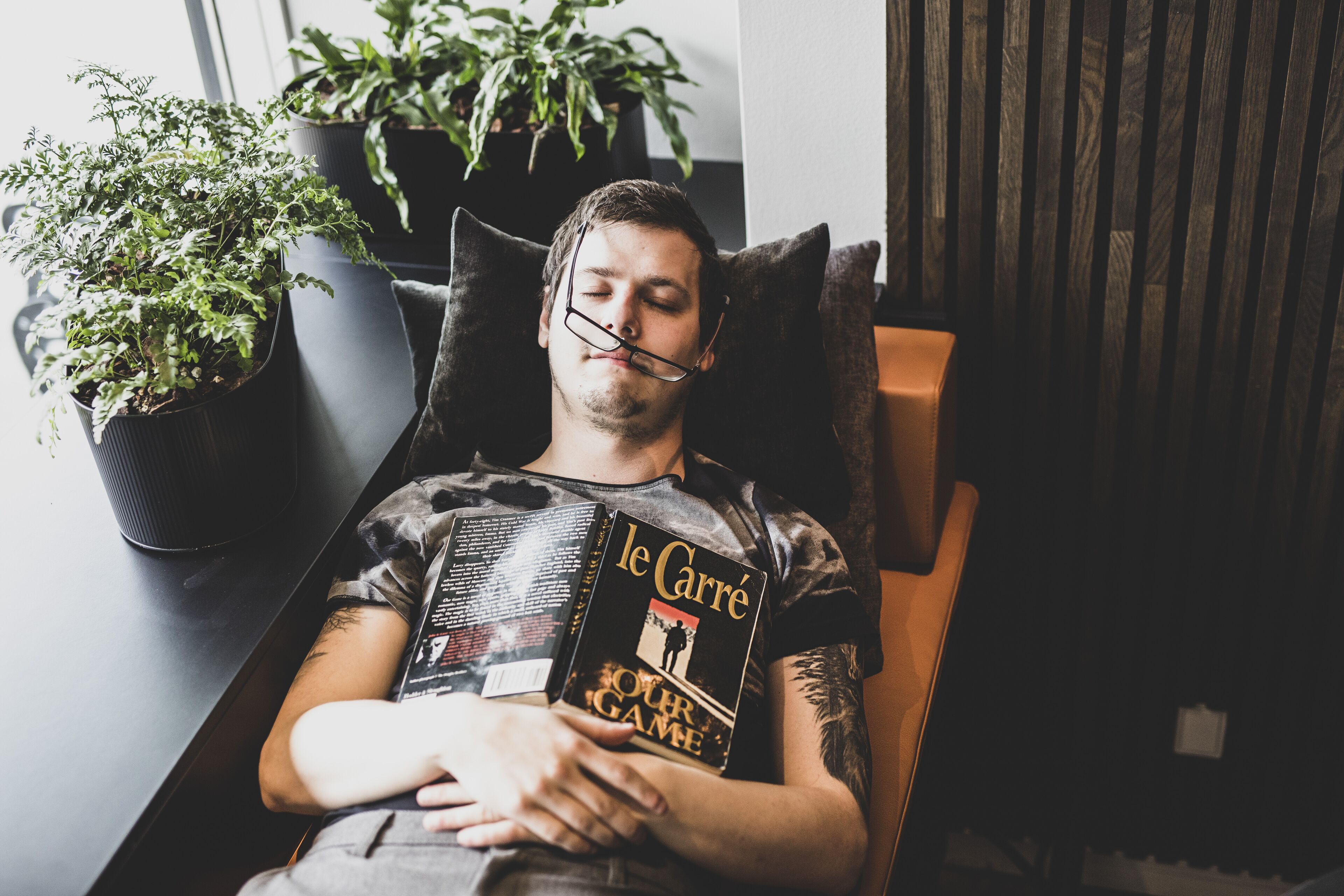 Una persona reclinada en una silla con un libro titulado 'Le Carré' y un tatuaje en el brazo.