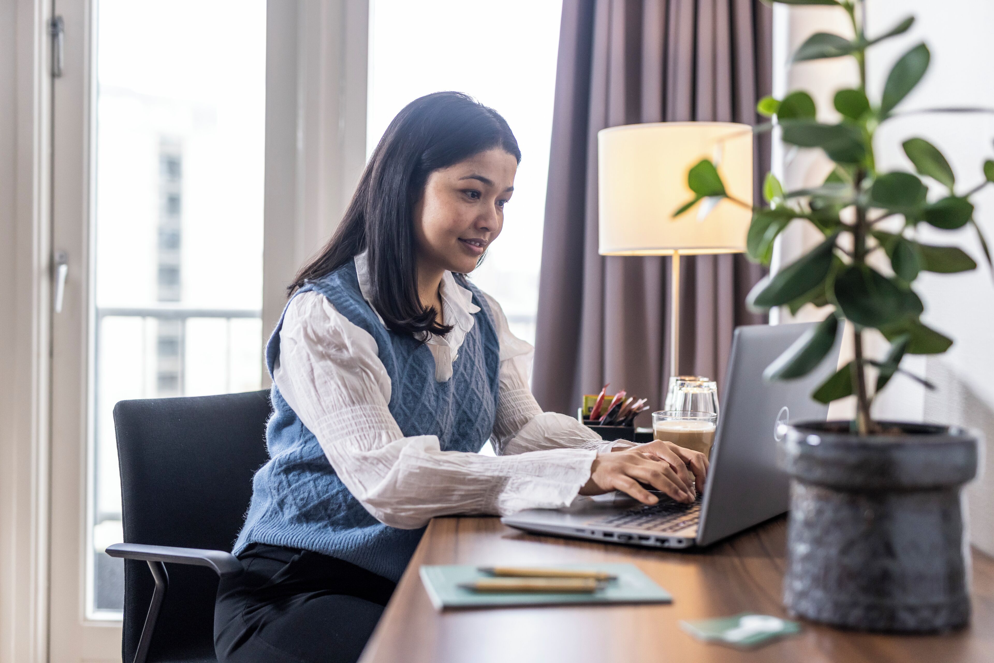 Une personne travaille sur un ordinateur portable, assise à un bureau éclairé par une lampe, avec une plante en pot et un verre d’eau à proximité.