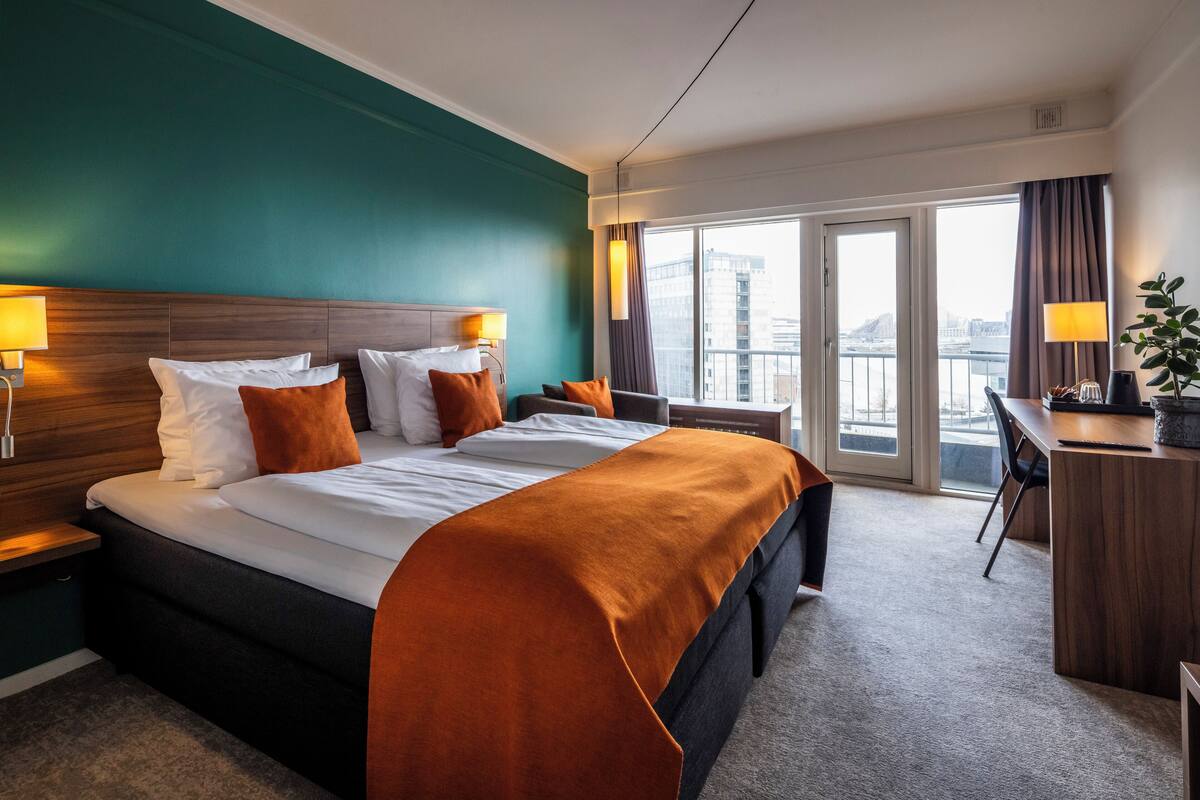 Et hotelværelse med en stor seng, et skrivebord og udsigt over byen fra vinduet.