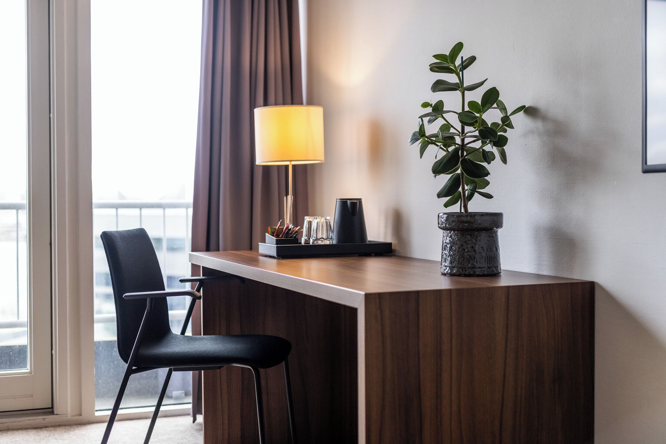 Een hotelkamer met een houten bureau, een zwarte stoel, een plant in pot, een lamp en een koffiezetapparaat.