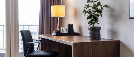 Une chambre d’hôtel avec un bureau en bois, une chaise noire, une plante en pot, une lampe et une machine à café.