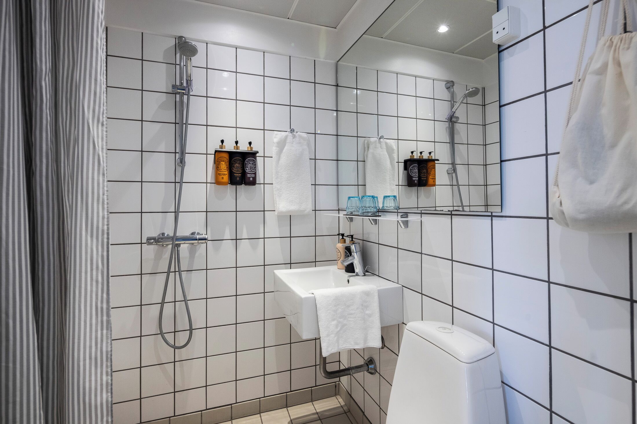 Een moderne badkamer met een witte wastafel, een spiegel en een douche. Er staan toiletartikelen op de muur en een handdoek hangt aan de wand.