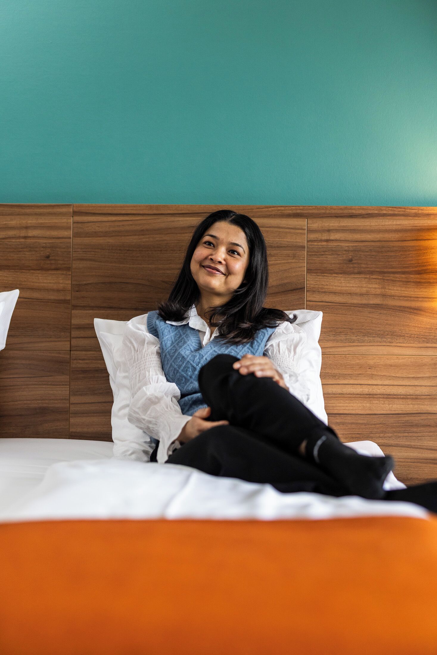 Una persona sentada en una cama con cabecera de madera, almohada blanca y manta naranja.