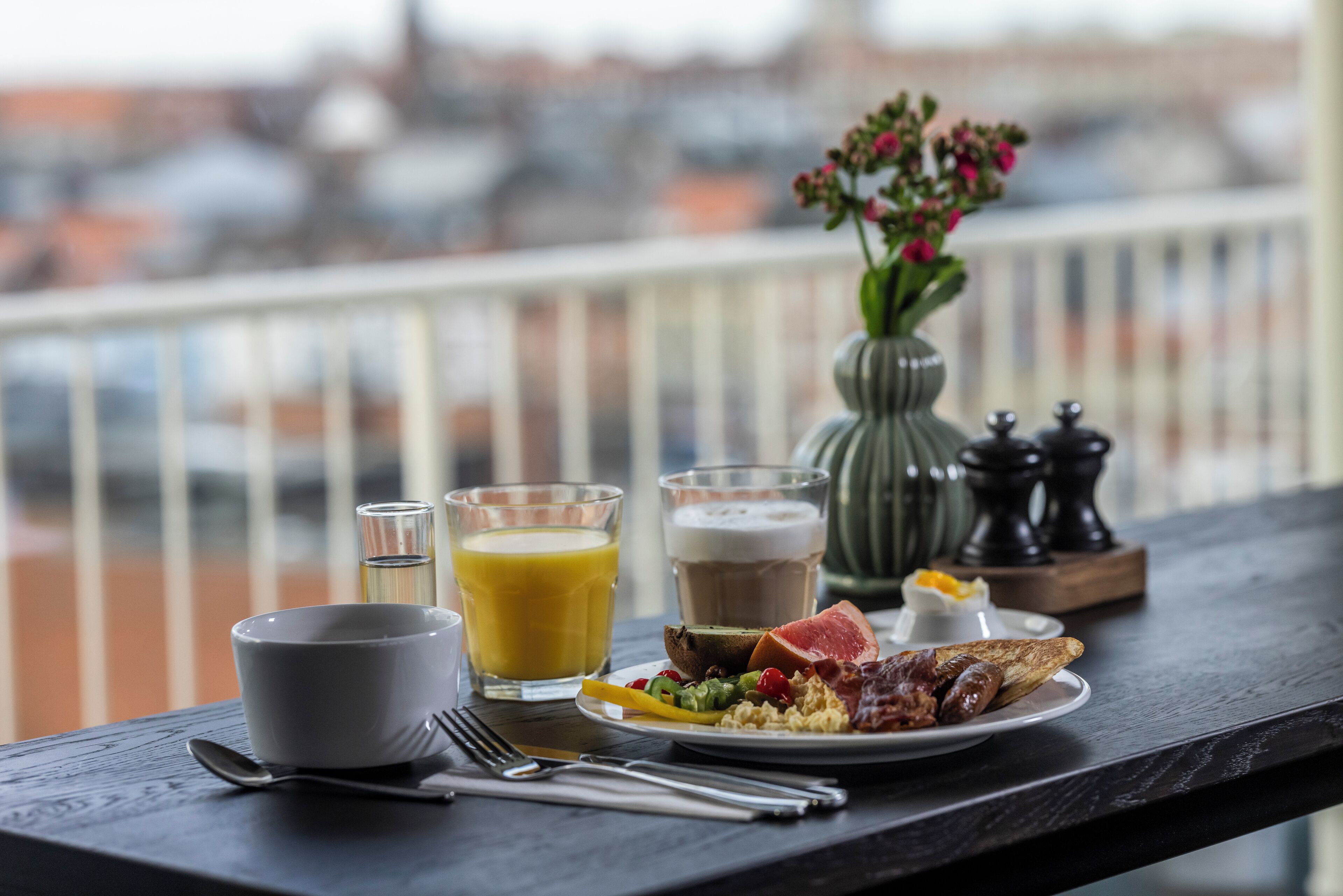A breakfast table with a plate of food, two glasses of juice, a cup of coffee, and a vase of flowers on a balcony overlooking a cityscape.