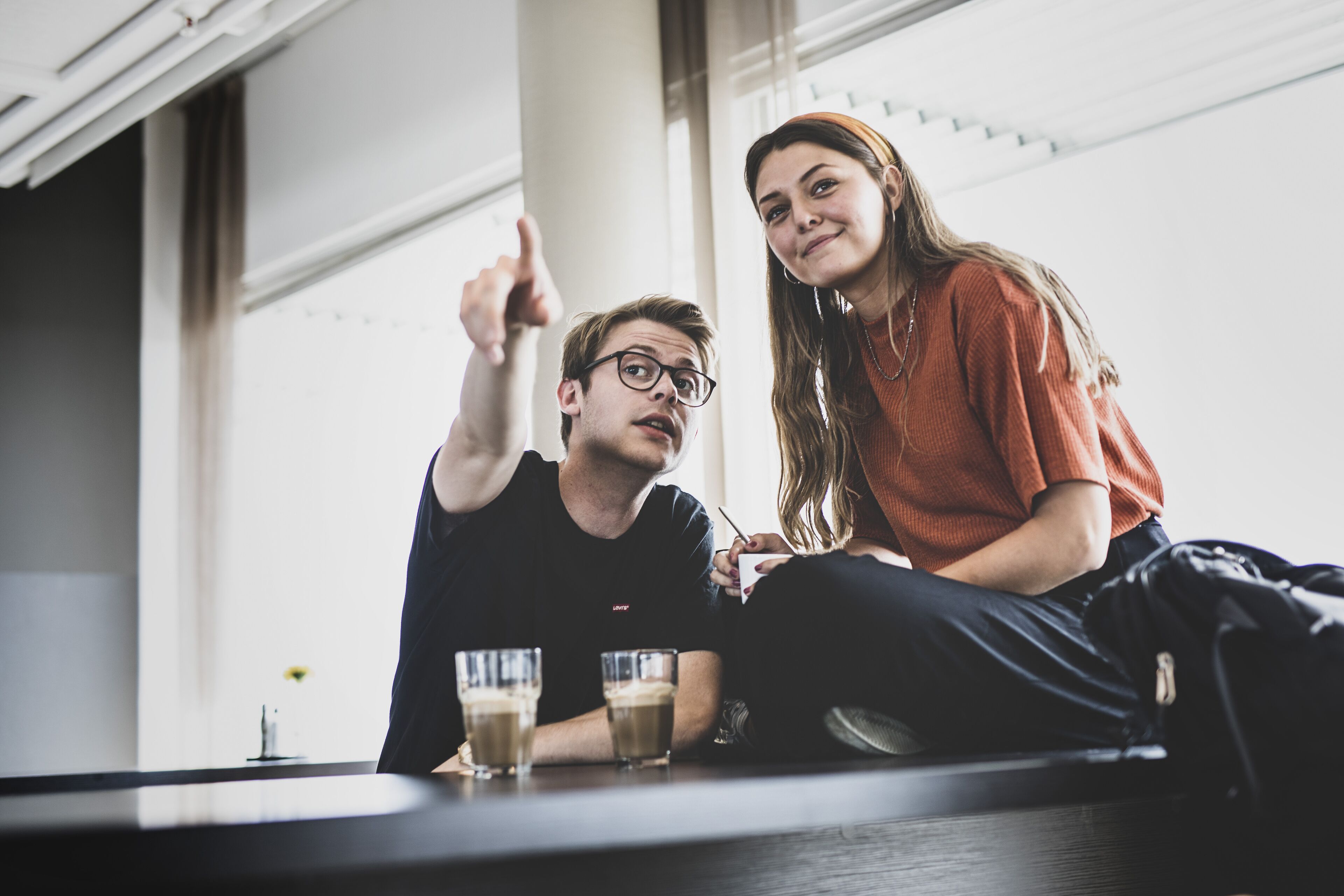 Deux personnes sont assises à une table, devant des boissons, dans une pièce dotée de grandes fenêtres et présentant un design moderne.