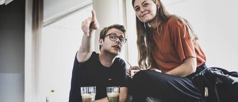 Deux personnes sont assises à une table, devant des boissons, dans une pièce dotée de grandes fenêtres et présentant un design moderne.