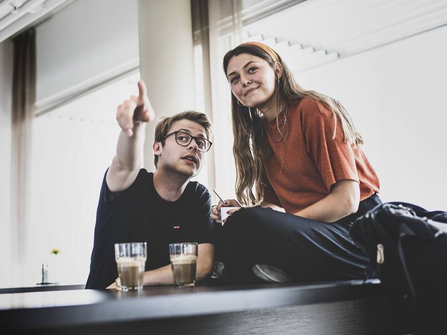 Dos personas sentadas en una mesa con bebidas, en una habitación con amplios ventanales y diseño moderno.