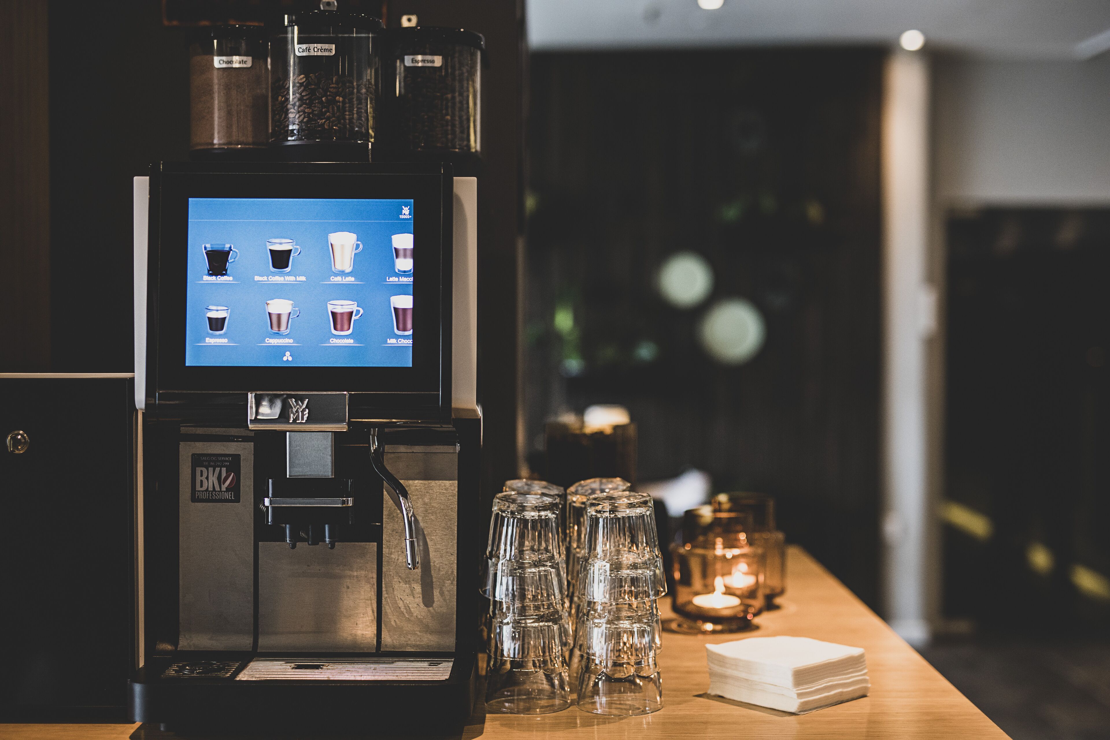 A coffee machine with a digital display menu, stacked glasses, and a candle on a wooden table.