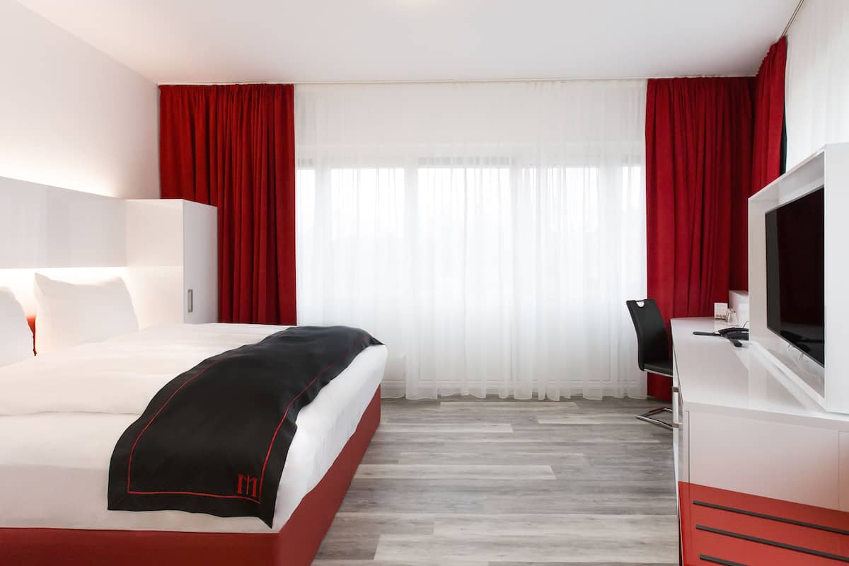 Habitación de hotel con cama, escritorio con silla, televisión y una ventana grande con cortinas rojas.