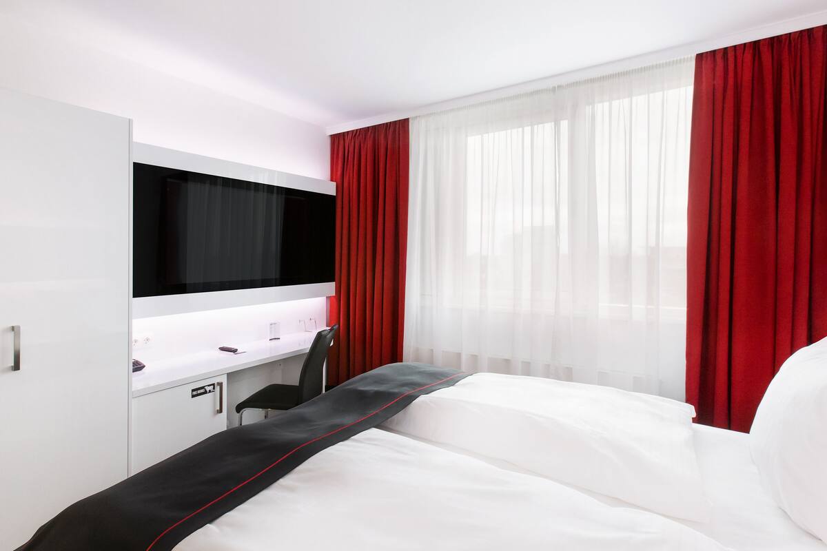 Una habitación de hotel con cama, escritorio, silla, televisor y cortinas rojas.