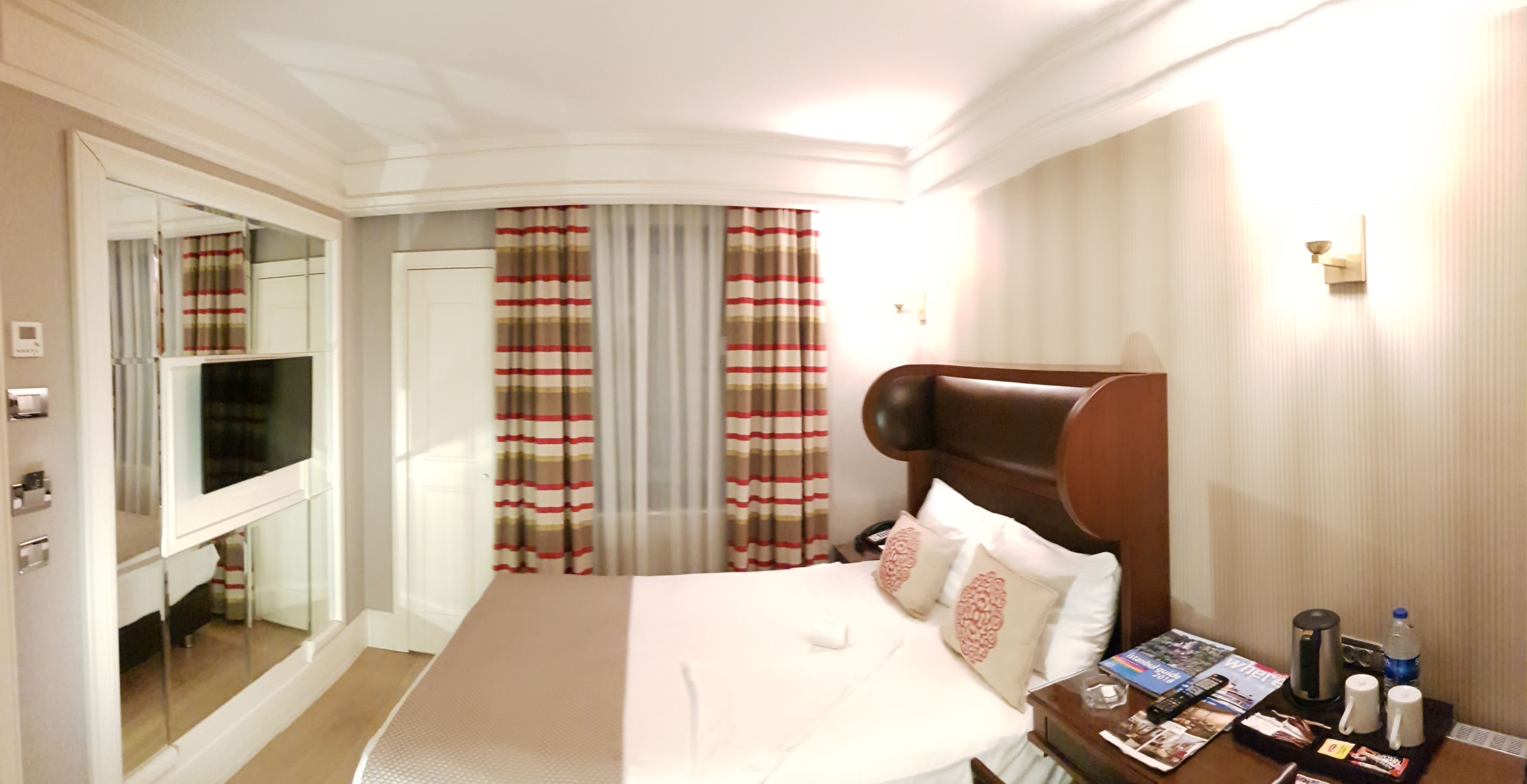 A hotel room with a large bed, a bedside table with a magazine and a coffee maker, and a mirror on the wall.