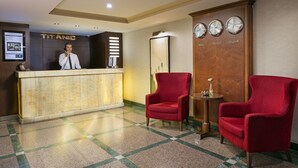 A hotel reception area with a marble counter, a receptionist, and two red armchairs.