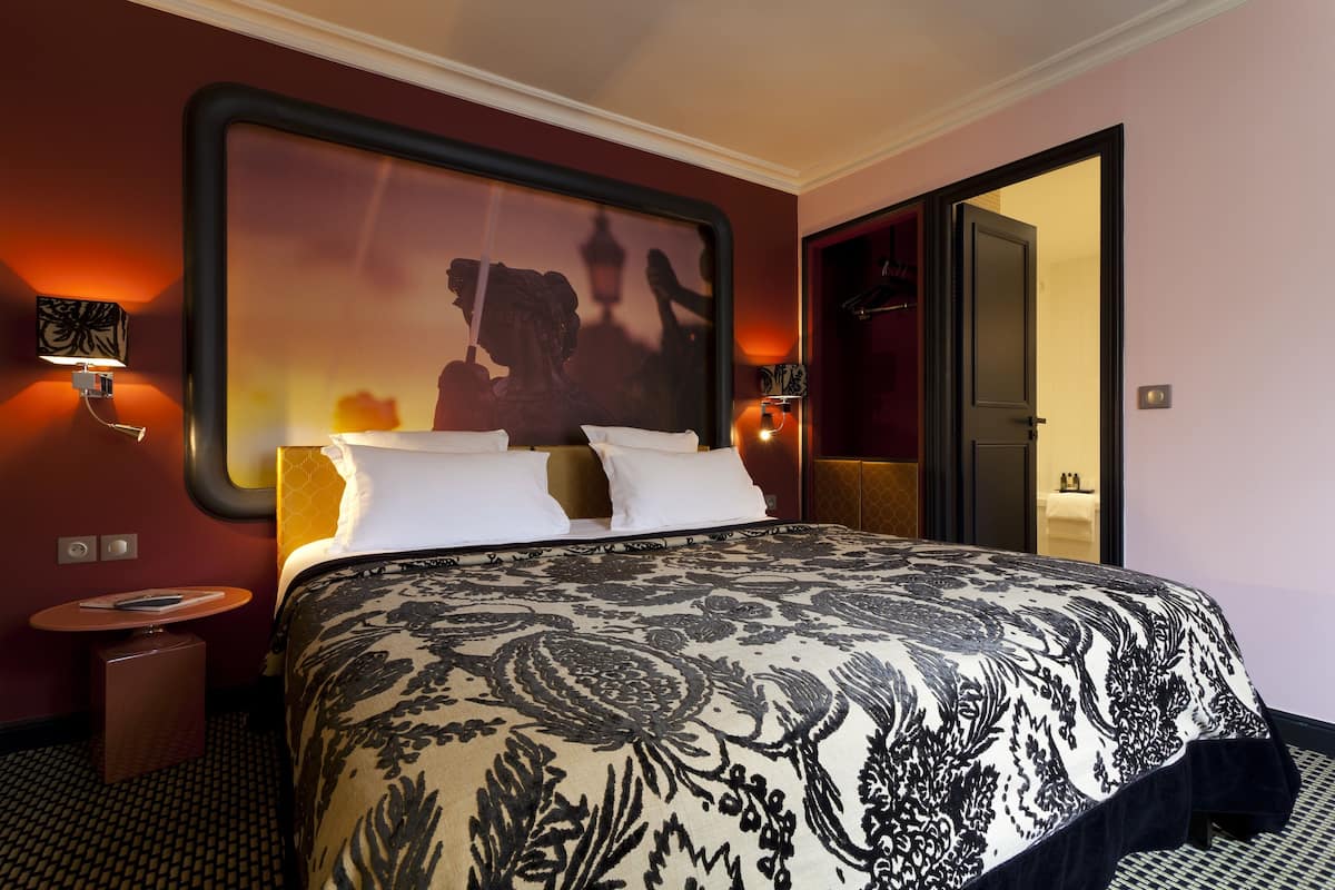Een hotelkamer met een groot bed, nachtkastje, wandlamp en een spiegel die een beeld weerkaatst.