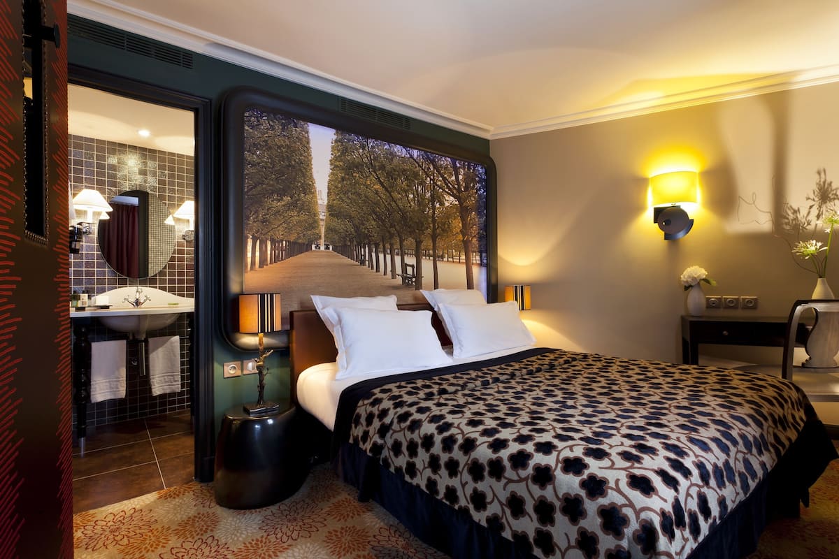 Een hotelkamer met een groot bed, een badkamer met een wastafel en spiegel, en uitzicht op bomen door een raam.