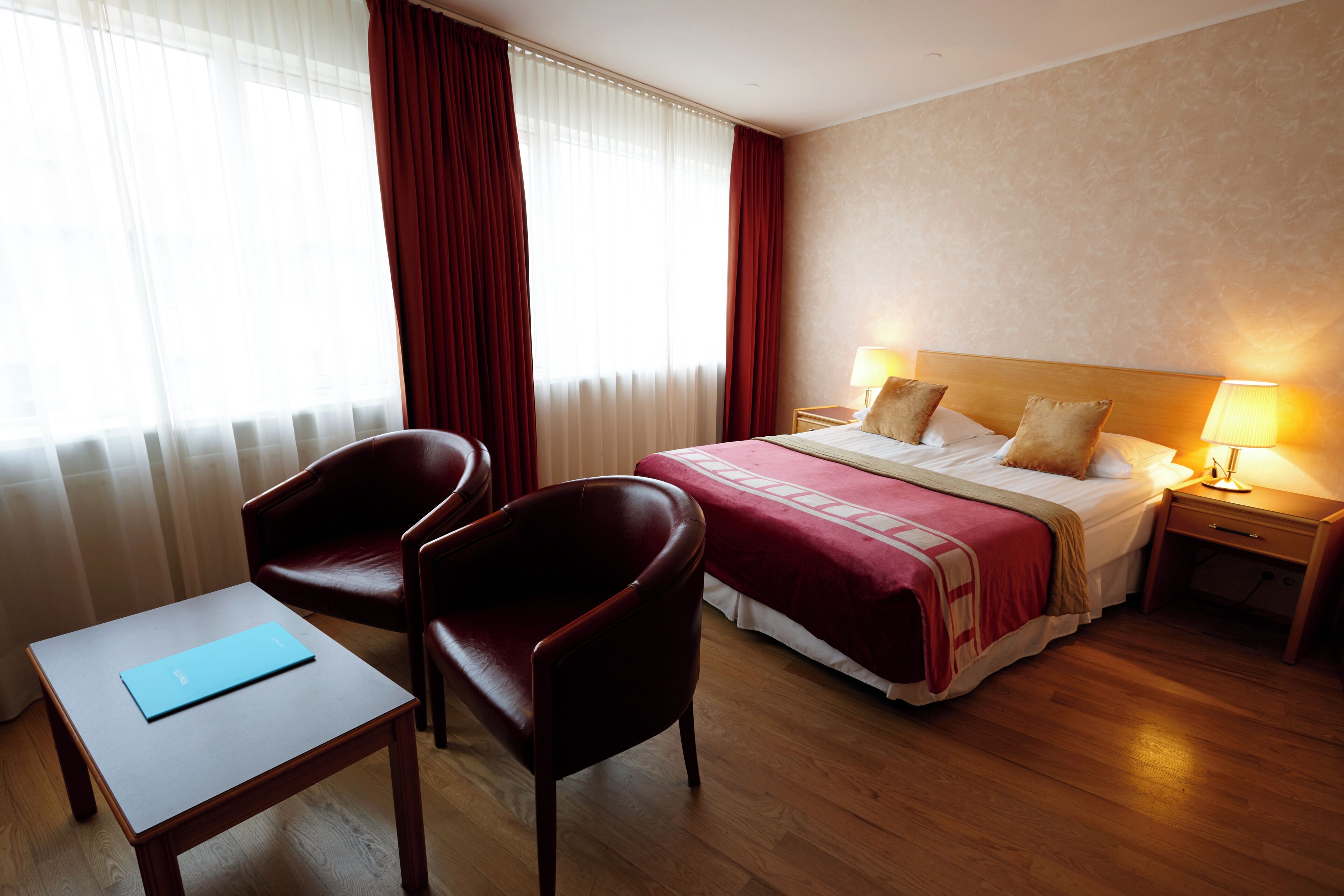 Habitación de hotel con cama, dos sillas, una mesita, una mesita de noche con lámpara y ventana con cortinas.