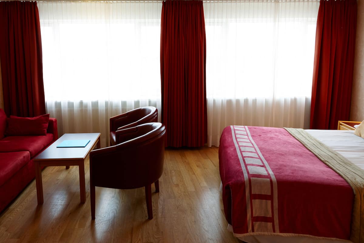 Ein Hotelzimmer mit rotem Sofa, kleinem Tisch, Stuhl, Bett mit rotem und weißen Überwurf und Fenster mit transparenten Vorhängen.