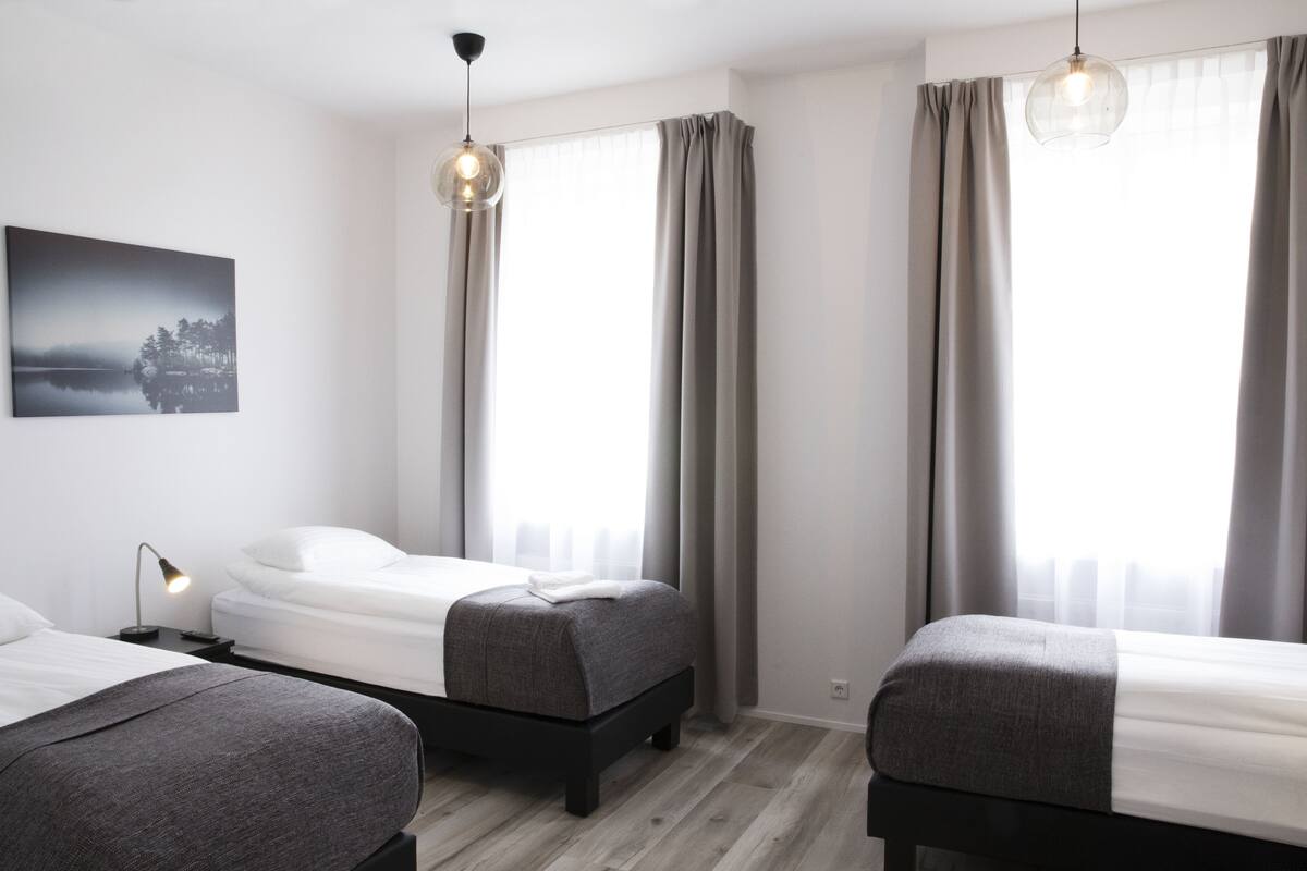 Ein modernes Hotelzimmer mit zwei Betten, einem Nachttisch mit Lampe, einem großen Fenster mit Vorhängen und einer Pendelleuchte.