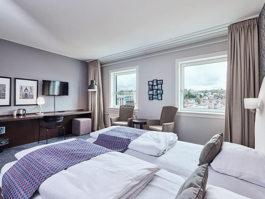 A hotel room with two beds, a desk, a chair, a TV, and a view of the city.