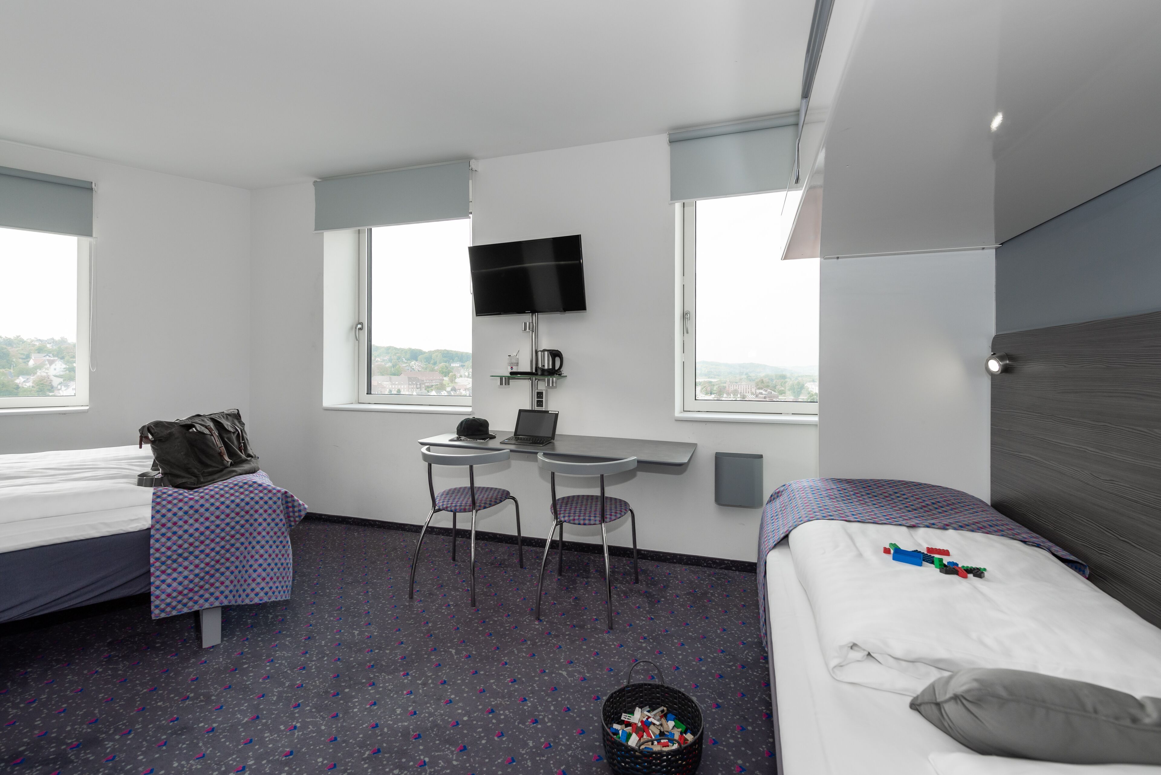 Habitación de hotel con dos camas, un escritorio con una computadora portátil, un televisor y una ventana con vista a la ciudad.