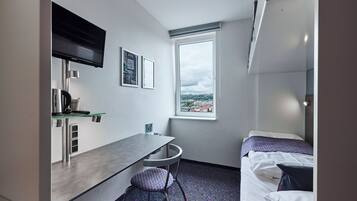 Une chambre d’hôtel compacte comprenant un coin cuisine, un lit simple, une petite table et une fenêtre donnant sur la ville.