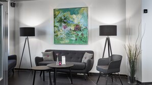 Un espace d’attente moderne avec un canapé gris, deux fauteuils, une table basse et deux lampadaires. Un grand tableau abstrait orne le mur.