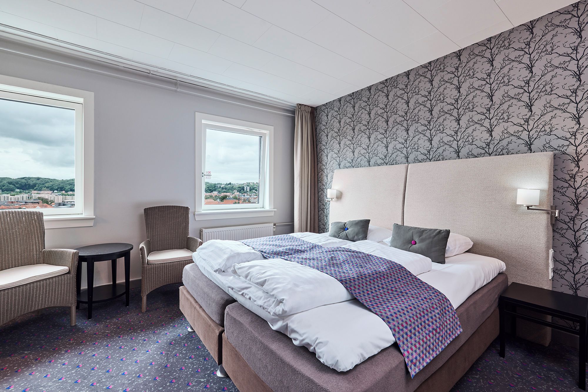 Habitación de hotel con una cama grande, una silla, una mesita y vistas a la ciudad.