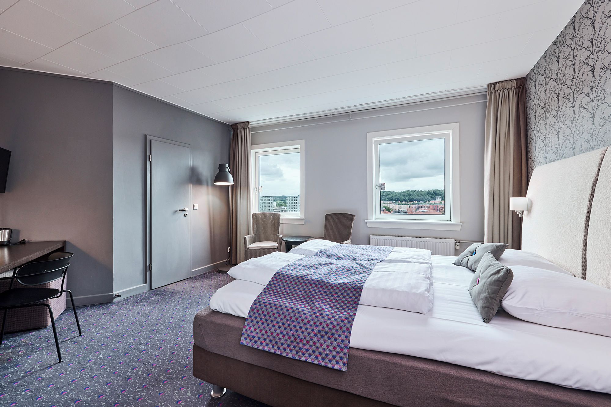 A hotel room with a bed, a desk, a chair, and a view of the city.