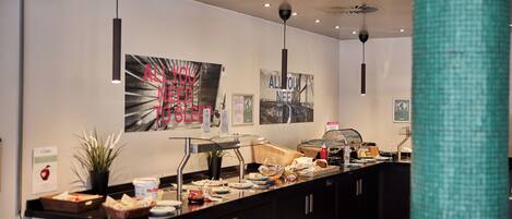 Zona de desayuno tipo buffet con diversas opciones de comida, una estación de café y una pared con carteles motivacionales.