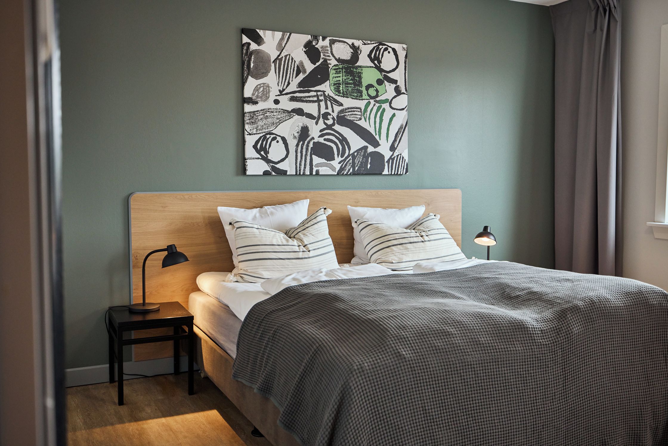 A bedroom with a bed, pillows, a bedside lamp, a small table, and a wall art piece.