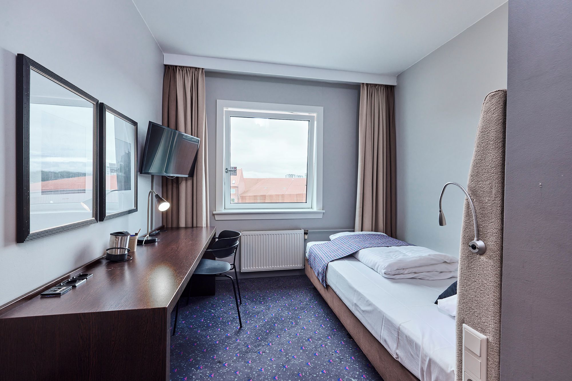 A hotel room with a bed, a desk with a computer, a TV, and a window with curtains.