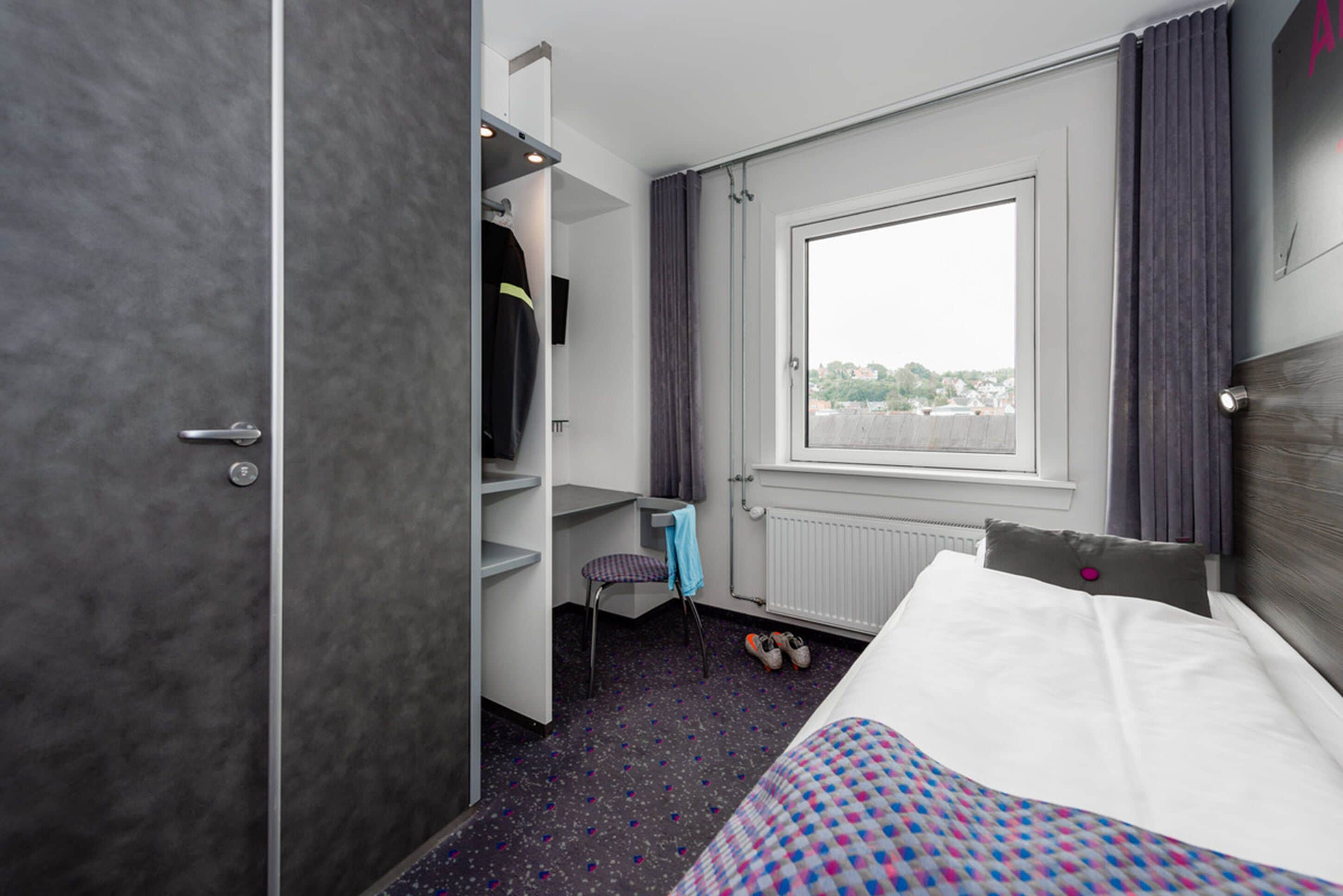 Una habitación de hotel con una cama, una silla, un escritorio y un ventanal con cortinas.