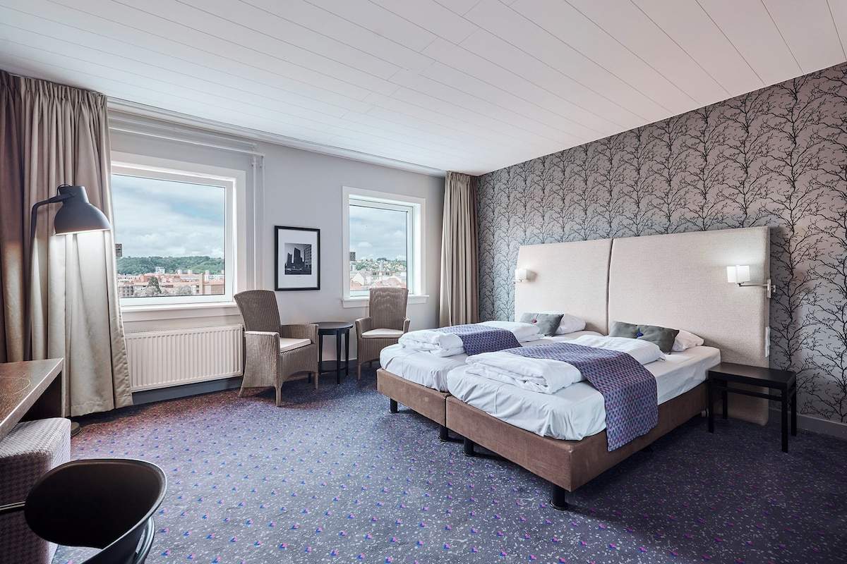 A hotel room with a large bed, two chairs, a desk, and a view of the city.
