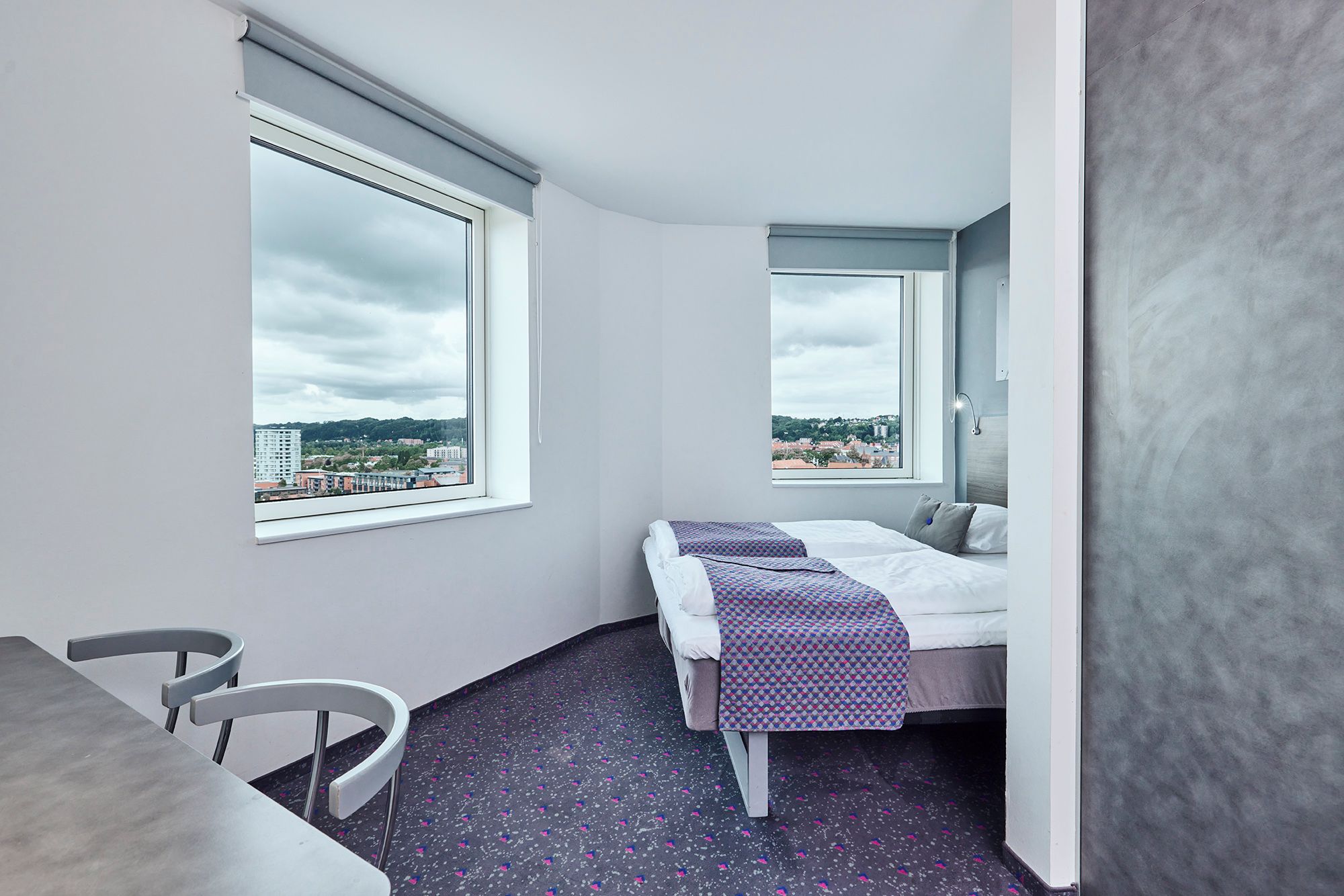 Habitación de hotel con cama, una silla, una mesa y vistas de la ciudad a través de amplios ventanales.