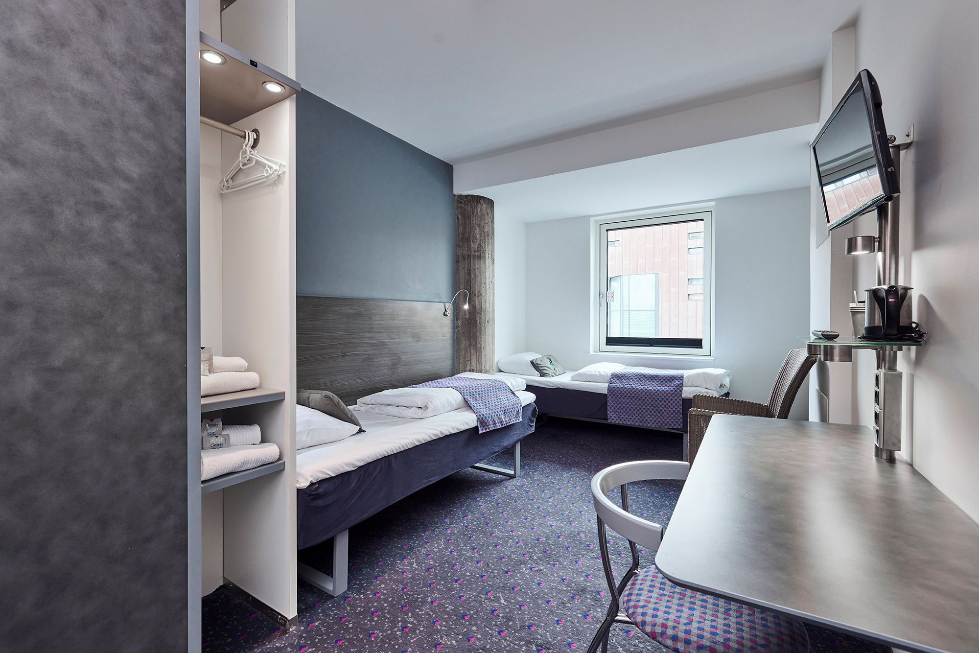 A hotel room with two beds, a desk, a chair, a TV, and a window with a view of buildings.