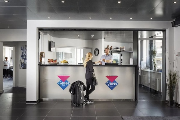 A modern hotel reception area with a glass partition, a digital clock, and a counter displaying food items.