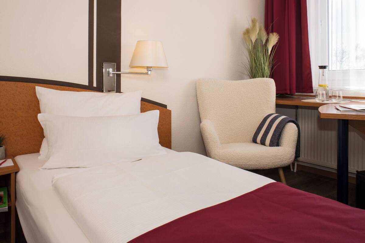 Une chambre d’hôtel comprenant un lit, une chaise, une table de chevet, une lampe, une fenêtre avec des rideaux et un radiateur.