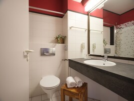 Un baño moderno con inodoro blanco, un lavabo con encimera oscura, un taburete de madera con toallas, un secamanos y un espejo con una pared de acento roja.