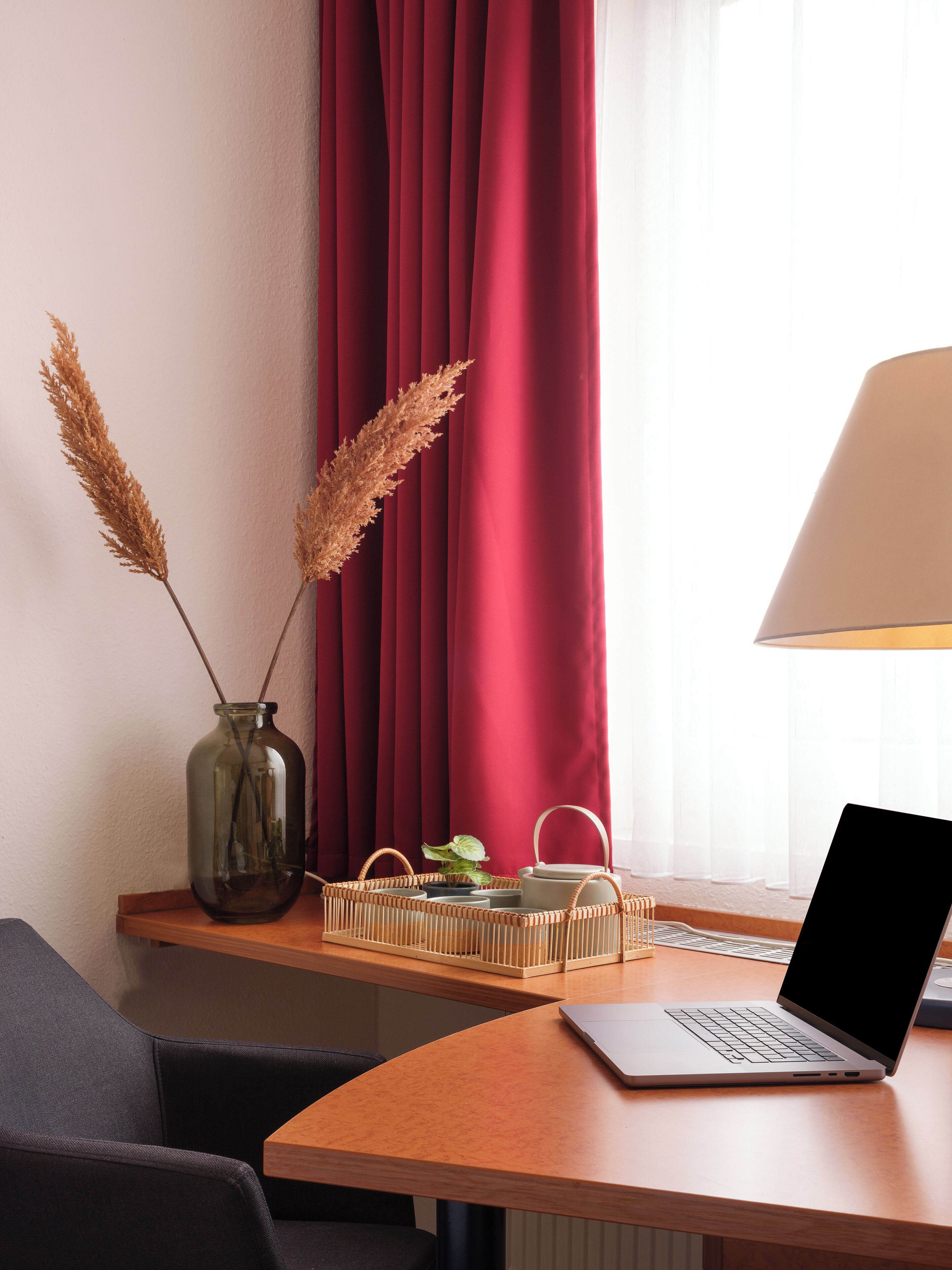 Ein Laptop auf einem Holztisch zusammen mit einer Vase mit getrocknetem Pampasgras, einem gewebten Korb und einer Lampe.